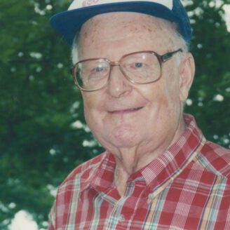 An older gentleman wearing glasses and a baseball cap smiles at the camera.