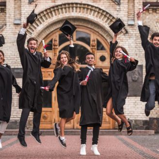 A group of six young people wearing caps and gowns jump in the air to celebrate graduation.