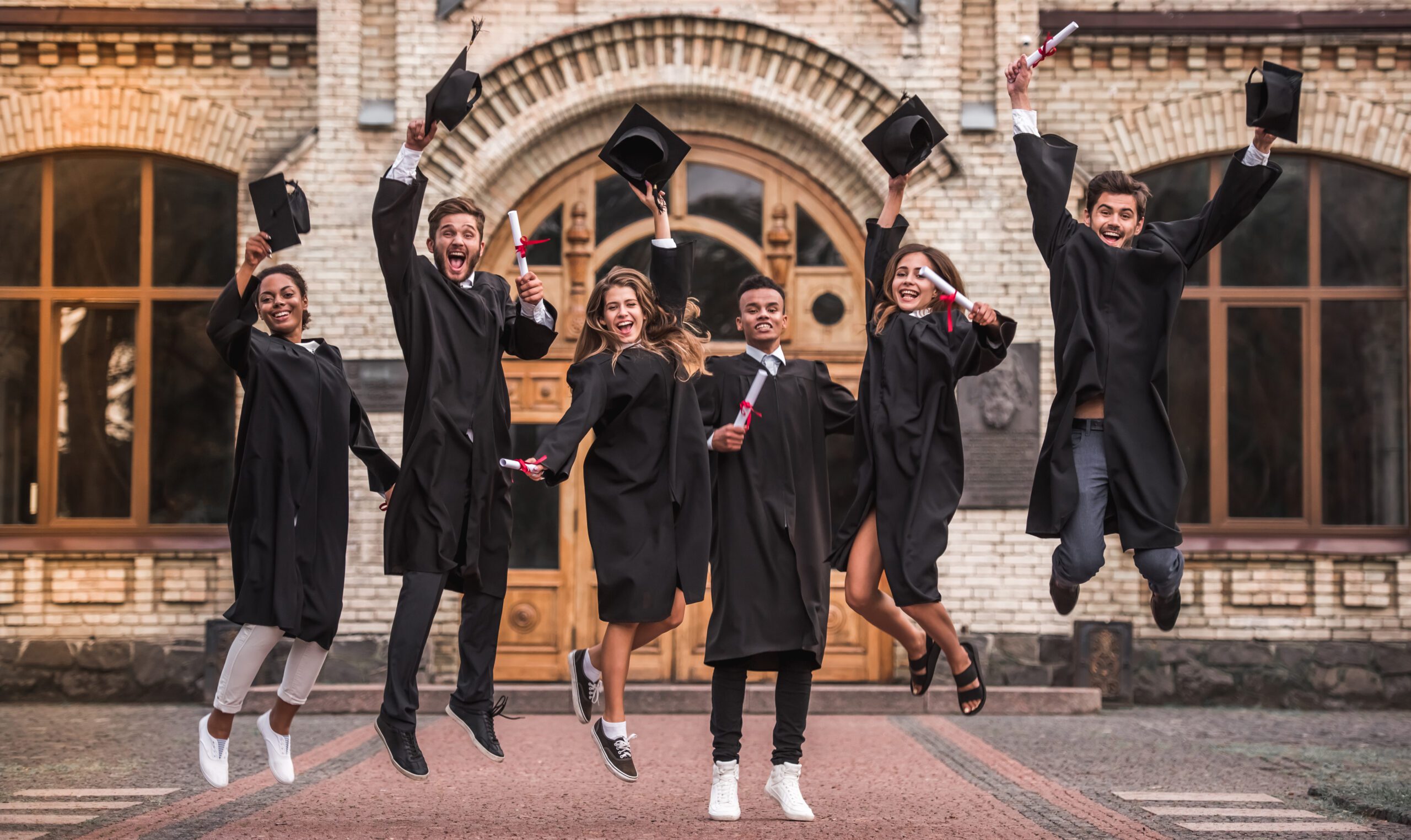 A group of six young people wearing caps and gowns jump in the air to celebrate graduation.