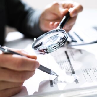 A close up of hands holding a pen and magnifying glass looking at a financial document.