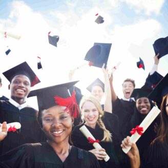 A group of young adults wearing graduation gowns smile at the camera and throw diplomas and caps into the air.