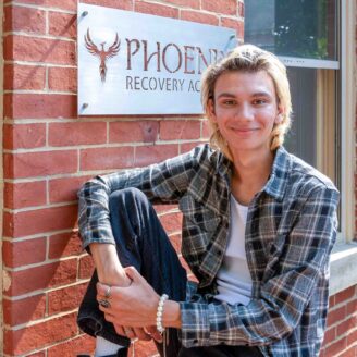 A young man sits on a brick porch in front of a sign that reads "Phoenix Recovery Academy".