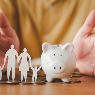 Hands form around a piggy bank next to a cutout of a family and coins.
