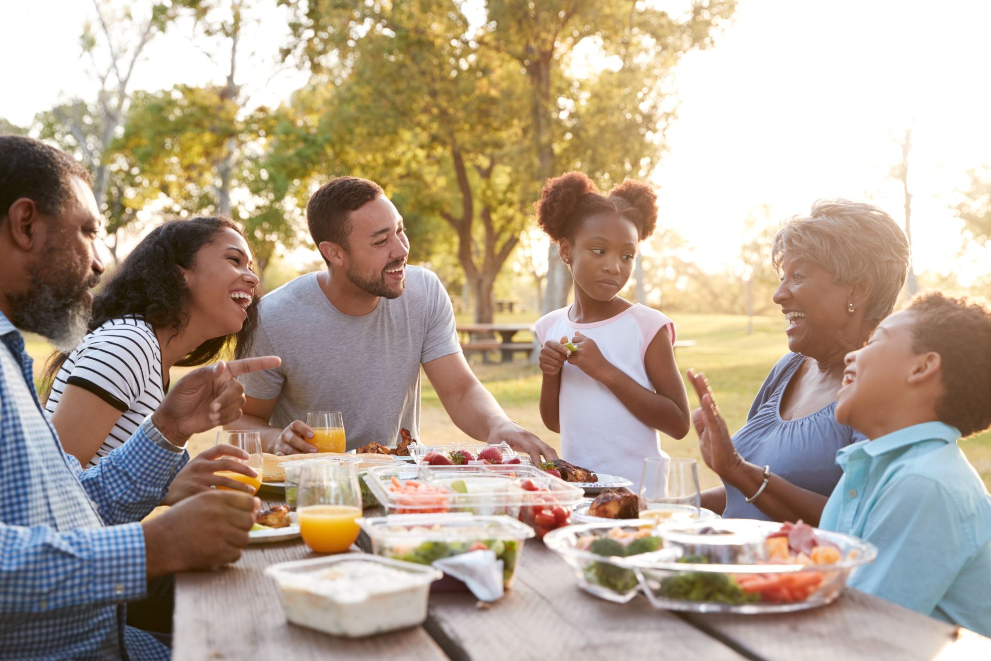 A multi-generational family laughs around a picnic table with healthy food. 