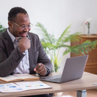 African American Businessman sits at a laptop laughing with paperwork on the table in front of him