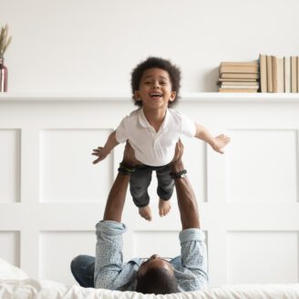 Black child laughing being held in the air over a parent laying in bed.