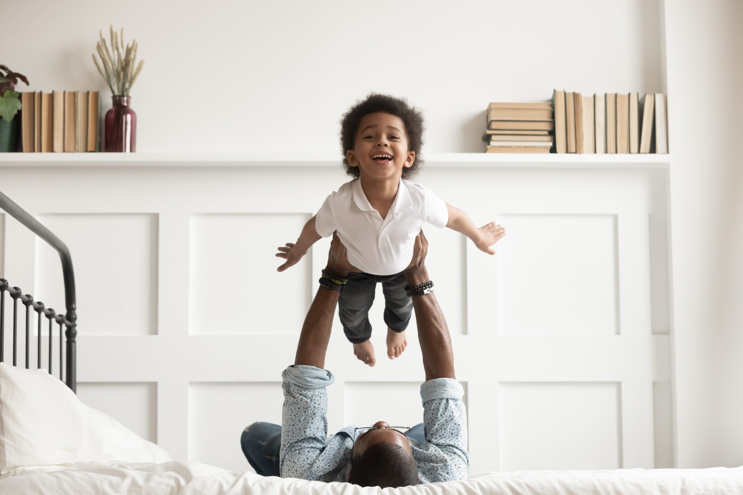 Black child laughing being held in the air over a parent laying in bed. 