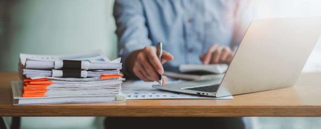 person at a desk working on a laptop with a stack of papers next to them