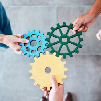 Three hands holding gears of blue, green, and yellow.