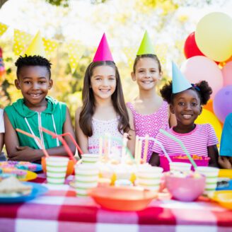 A group of children sit around a table smiling. They are surrounded by balloons and other birthday party decor.