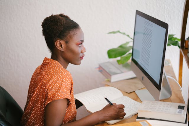 woman looks at a computer screen while writing in a notebook at her desk