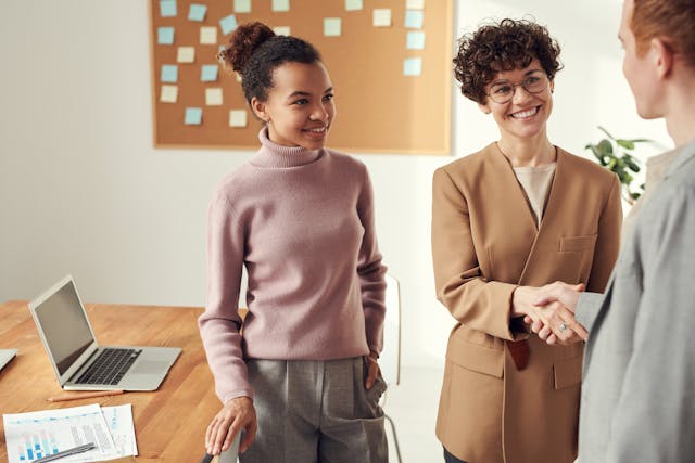 two women smiling one is shaking hands with a third woman. They are standing in an office
